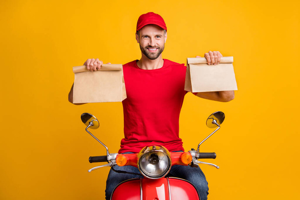 Photo of cheerful guy ride bike hold two paper packages wear red t-shirt headwear isolated yellow color background - Fotoğraf, Görsel