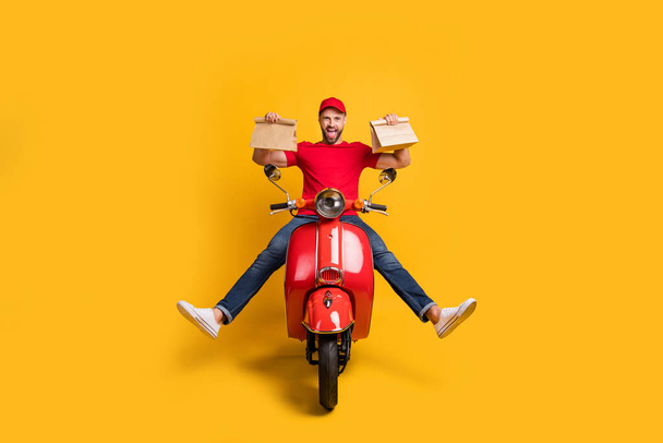 Photo of careless crazy guy drive bike hold two packages wear red t-shirt cap jeans isolated yellow color background - Foto, Imagem