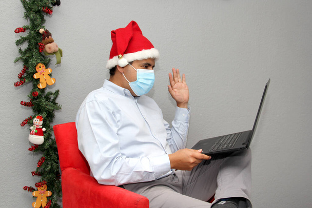 Koruyucu maskeli Latin bir adam, video konferansında Noel Baba şapkası ve laptop, Noel süslemesi, yeni normal Cvid-19. - Fotoğraf, Görsel