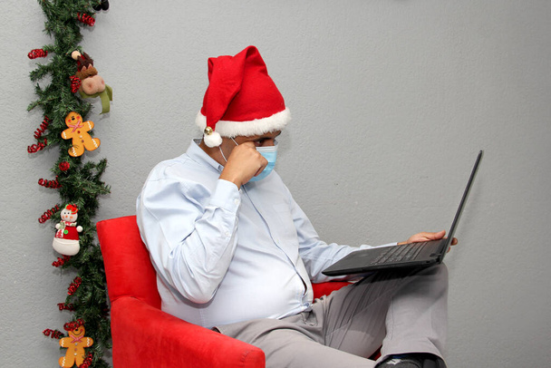 lateinischer Mann mit Schutzmaske, Weihnachtsmann-Hut und Laptop in Videokonferenz, Weihnachtsdekoration, neue normale covid-19 - Foto, Bild
