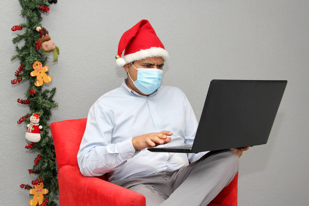 Koruyucu maskeli Latin bir adam, video konferansında Noel Baba şapkası ve laptop, Noel süslemesi, yeni normal Cvid-19. - Fotoğraf, Görsel