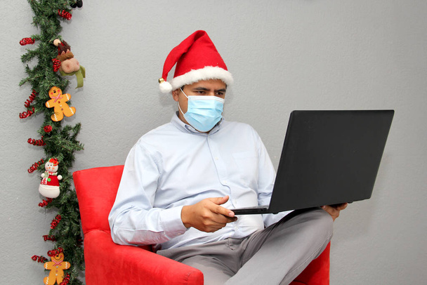 latinský muž s ochrannou maskou, čepicí Santa Claus a notebookem ve videokonferenci, vánoční ozdoba, nová normální covid-19 - Fotografie, Obrázek