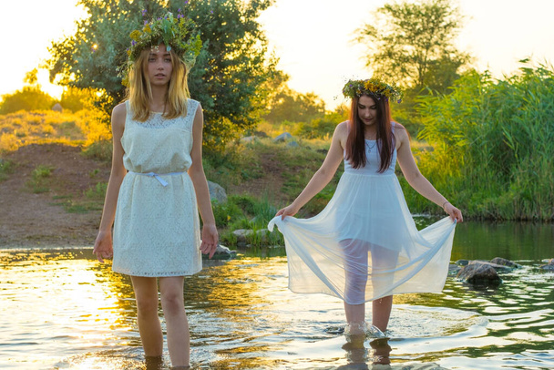 duas namoradas, meninas em vestidos brancos e coroas de flores em suas cabeças, de pé em um rio de água, perto da costa, rindo, contra o pôr do sol. O conceito de virgindade, beleza natural, meninas de vinte anos de pé pés descalços em th - Foto, Imagem
