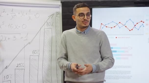 Mittlere Nahaufnahme eines smarten Geschäftsmannes aus dem Nahen Osten, der vor Mitarbeitern spricht und Details eines neuen Projekts anhand eines gezeichneten Diagramms mit Zahlen auf einem Flipchart erklärt - Filmmaterial, Video