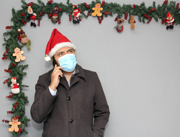 Latinský muž s ochrannou maskou, kabátem a čepicí Santa Claus ve videohovoru s mobilním telefonem a vánoční výzdobou, nový normální covid-19 - Fotografie, Obrázek
