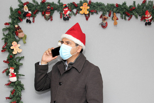 Homme latin avec masque de protection, manteau et chapeau du Père Noël en appel vidéo avec téléphone portable et décoration de Noël, nouveau covid-19 normal - Photo, image