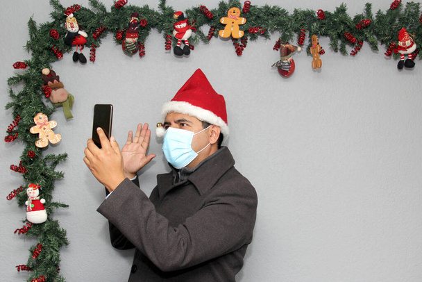 Latinalainen mies, jolla on suojanaamari, takki ja joulupukin hattu videopuhelussa matkapuhelimella ja joulukoristeella, uusi normaali covid-19 - Valokuva, kuva