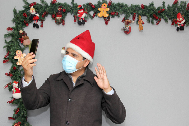 Latin man with protection mask, coat and Santa Claus hat in video call with cell phone and Christmas decoration, new normal covid-19 - Photo, Image