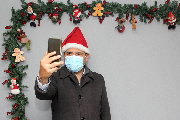 Homme latin avec masque de protection, manteau et chapeau du Père Noël en appel vidéo avec téléphone portable et décoration de Noël, nouveau covid-19 normal - Photo, image