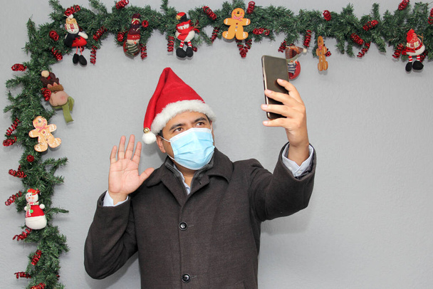 Koruyucu maskeli, ceketli ve Noel Baba şapkalı Latin bir adam cep telefonu ve Noel süslemesi ile görüntülü görüşme yapıyor. - Fotoğraf, Görsel