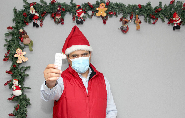 Hombre latino con máscara de protección, chaleco rojo y sombrero de Papá Noel, con medicina y decoración navideña, nuevo covid-19 normal - Foto, Imagen