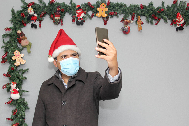 Koruyucu maskeli, ceketli ve Noel Baba şapkalı Latin bir adam cep telefonu ve Noel süslemesi ile görüntülü görüşme yapıyor. - Fotoğraf, Görsel