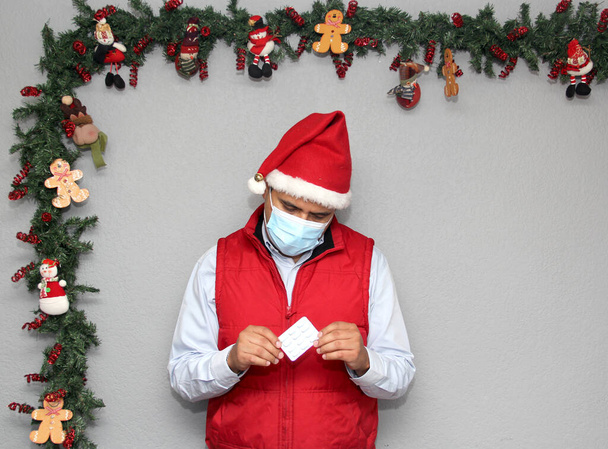 Hombre latino con máscara de protección, chaleco rojo y sombrero de Papá Noel, con medicina y decoración navideña, nuevo covid-19 normal - Foto, Imagen