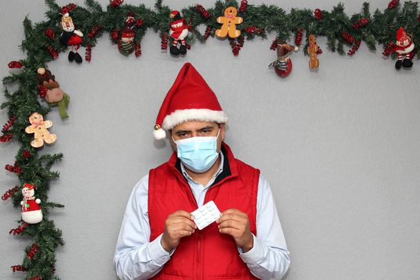 Homme latin avec masque de protection, gilet rouge et chapeau du Père Noël, avec médecine et décoration de Noël, nouveau covide normal-19 - Photo, image