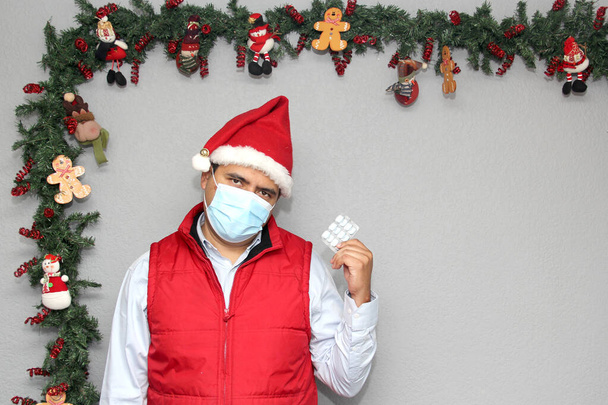 Korunma maskeli Latin bir adam, kırmızı yelek ve Noel Baba şapkalı, ilaç ve Noel süslemeli, yeni normal CVid-19. - Fotoğraf, Görsel