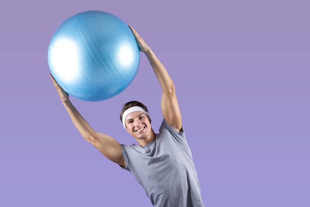 Positivo joven deportista levantando la pelota de fitness por encima de su cabeza, doblando a un lado sobre fondo de estudio lila, espacio de copia - Foto, imagen
