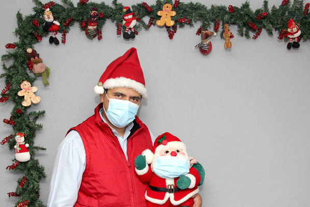 hombre latino con máscara de protección y muñeca de Santa Claus en la decoración de Navidad, nuevo covid-19 normal - Foto, imagen