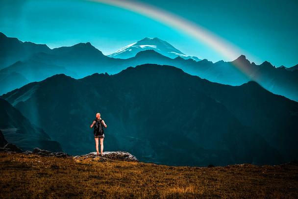 Girl traveler with backpack stands on a stone back to the viewer. She looks at the snow-capped top. Picturesque cloudless landscape with a rainbow. Wallpaper for concept of business thinking. sport - Zdjęcie, obraz