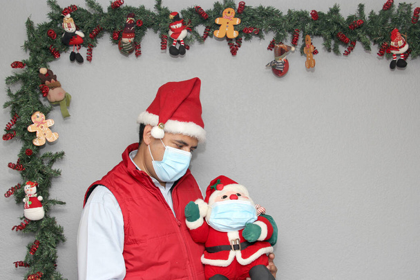 latinský muž s ochrannou maskou a Santa Claus panenkou v vánoční výzdobě, nový normální covid-19 - Fotografie, Obrázek