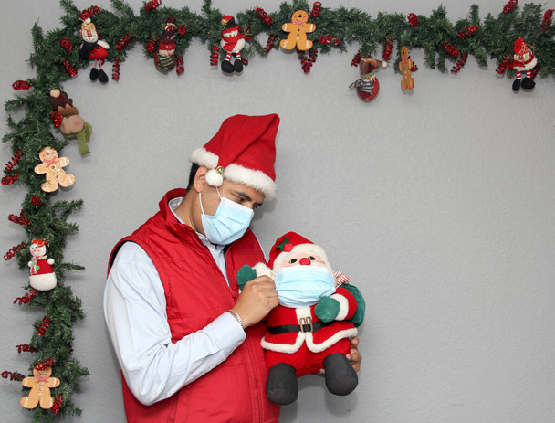hombre latino con máscara de protección y muñeca de Santa Claus en la decoración de Navidad, nuevo covid-19 normal - Foto, Imagen