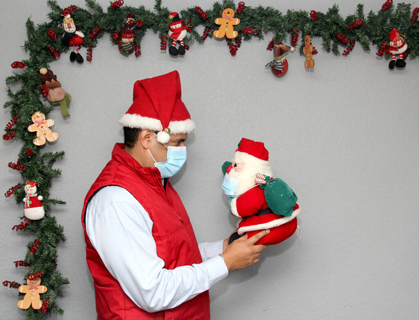 Koruyucu maskeli latin adam ve Noel Baba bebeği noel süslemesinde, yeni normal covid-19 - Fotoğraf, Görsel