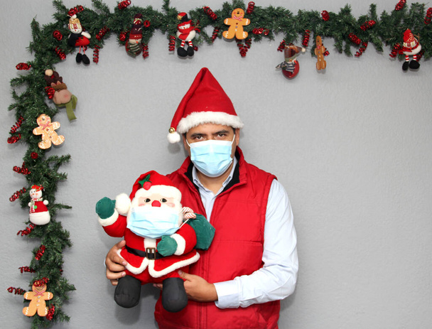 homme latin avec masque de protection et poupée santa claus dans la décoration de Noël, nouveau covid-19 normal - Photo, image