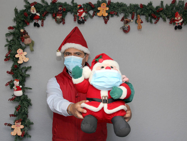 hombre latino con máscara de protección y muñeca de Santa Claus en la decoración de Navidad, nuevo covid-19 normal - Foto, Imagen