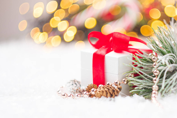 Kerstcadeau of cadeau met rood lint, kleine sparren of dennenappels, kralen, dennentak op de achtergrond van wazig licht van slinger. - Foto, afbeelding