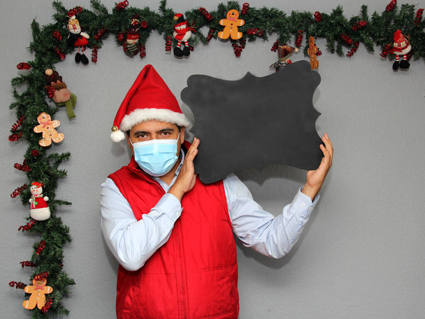 homem latino com máscara de proteção e quadro-negro para escrever mensagem, na decoração de Natal, novo normal covid-19 - Foto, Imagem