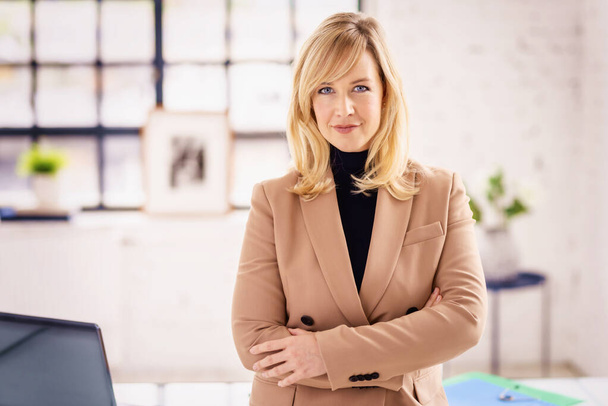 Potrait shot of beautiful blond haired businesswoman wearing turtleneck sweater and blazer while standing at the office with arms crossed.  - Fotoğraf, Görsel