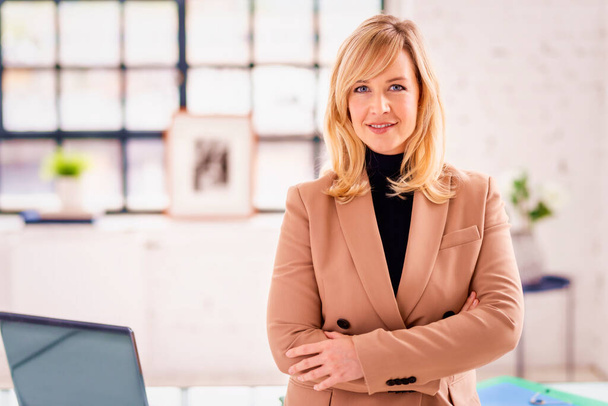 Potrait shot of beautiful blond haired businesswoman wearing turtleneck sweater and blazer while standing at the office with arms crossed.  - Foto, afbeelding