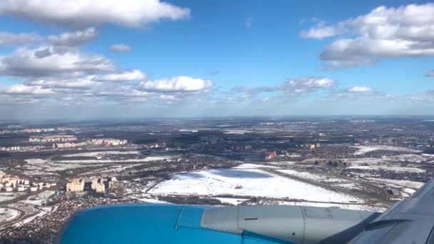 青い飛行機がモスクワの冬の街を飛んでいます。窓からの眺め:上から雲が立ち並ぶ青空、下からの住宅や道路 - 映像、動画