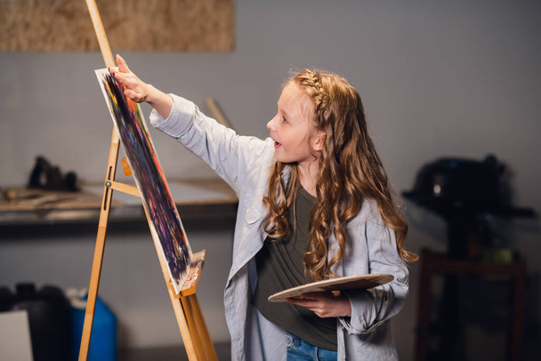 Νεαρή κοπέλα δημιουργικός καλλιτέχνης σε ένα εργαστήριο. Με πάθος ζωγραφίζει μια εικόνα βυθισμένη στη δουλειά. - Φωτογραφία, εικόνα