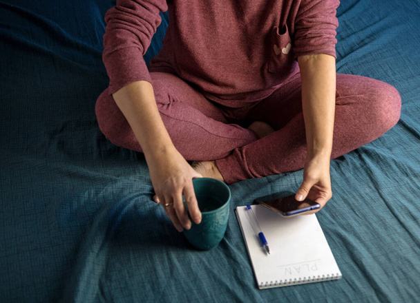 Kadın eşofmanla evde bir yatakta oturuyor. Notepad 'de gününü planla. Yatak çarşaflarının rengi tidewater yeşili, eğilim 2021 - Fotoğraf, Görsel