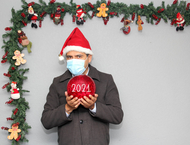 hombre latino con máscara de protección, sombrero de santa claus y esfera roja, decoración de Navidad, nuevo covid-19 normal - Foto, imagen