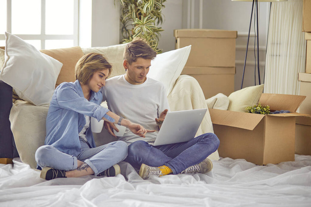 Portret szczęśliwej pary patrzącej razem na laptopa, siedzącej w nowym domu, otoczonej pudełkami - Zdjęcie, obraz