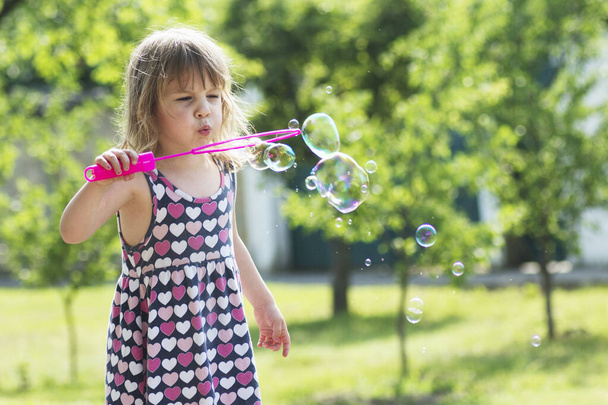 Милая маленькая девочка в платье раздувает мыльные пузыри на улице.. - Фото, изображение