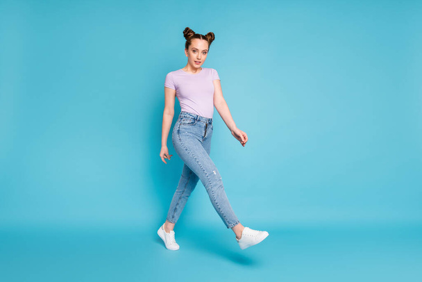 Full length profile photo of pretty cheerful lady two funny buns walking street good mood nice day weather wear casual purple t-shirt denim jeans shoes isolated blue color background - Fotó, kép