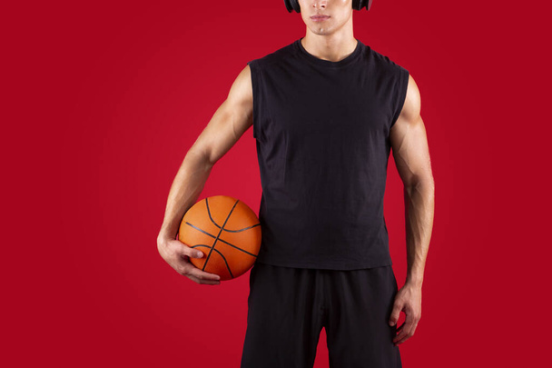 Cropped view of young basketball player holding ball, listening to music in headphones over red background, copy space - Zdjęcie, obraz