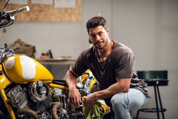 Красивый молодой мотоциклист в джинсах и футболке позирует для фото, сидящего на велосипеде в гараже - Фото, изображение