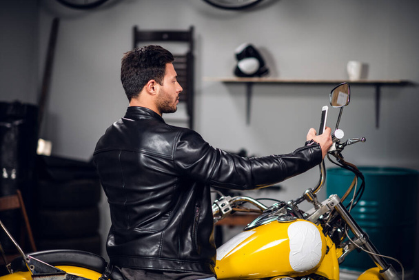 Ein schöner muskulöser Biker führt einen Videoblog und spricht über einen Videoanruf auf einem Smartphone, während er auf einem Motorrad in der Garage sitzt - Foto, Bild