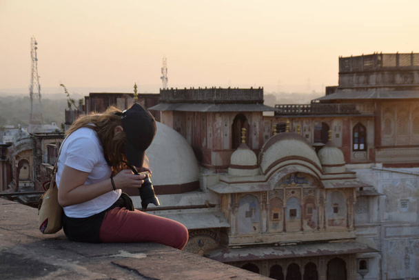 Podróżniczka i młoda kobieta naprawiają aparat, siedząc na dachu miasta. Piękny zachód słońca i panorama miasta w tle. Zdjęcie zrobione w Indiach. - Zdjęcie, obraz