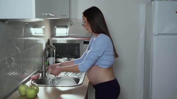 una donna incinta di aspetto europeo si trova in cucina a lavare un bicchiere. Lavorare duramente durante il concetto di gravidanza - Filmati, video