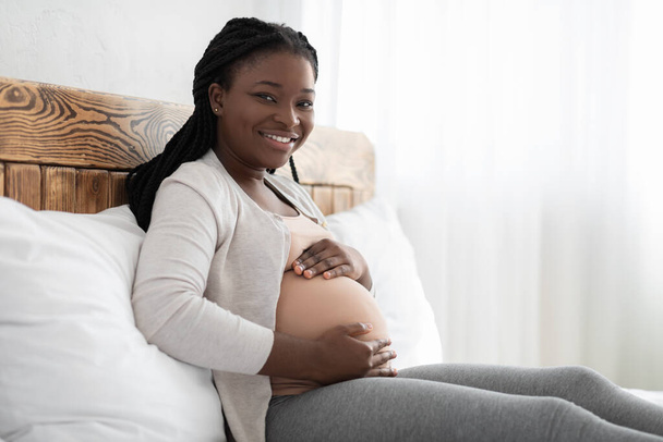 Усміхнена чорна вагітна жінка спить на ліжку і тримає руки на животі
 - Фото, зображення