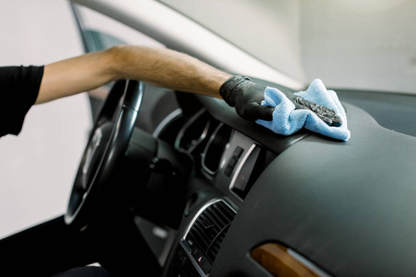 Cleaning the car, detailing concept, car wash. Close up cropped shot of hands of professional male worker, wearing rubber gloves, cleaning the interior of the car with microfiber cloth - Фото, изображение