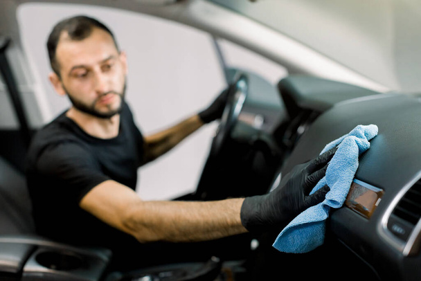 Cleaning service, car detailing concept. Young man, auto service worker, washing a car interior, console and dashboard with microfiber cloth, in a professional car wash. Fpcus on a hand with cloth - Foto, imagen