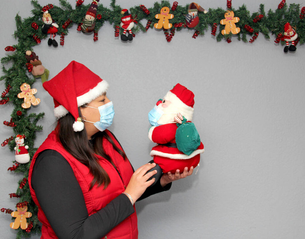 Λατινική γυναίκα με μάσκα προστασίας, καπέλο και Άη Βασίλη κούκλα στη διακόσμηση των Χριστουγέννων, νέα κανονική covid-19 - Φωτογραφία, εικόνα