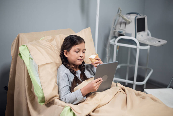 Позитивная маленькая девочка использует приложение на своем цифровом устройстве, находясь в больничной койке - Фото, изображение