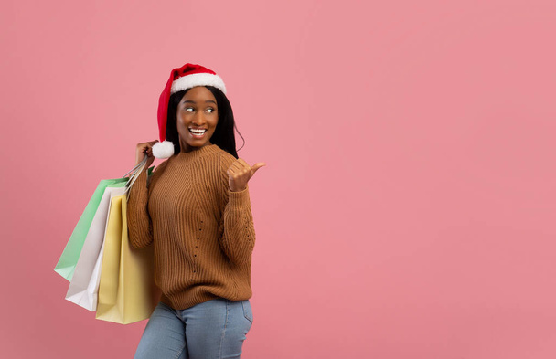 Huge Christmas sale. Attractive black woman in Santa hat holding gift bags and pointing at blank space, pink background - Foto, Imagen