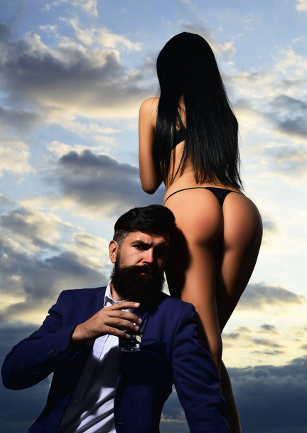 Bearded businessman in elegant suit with glass of whiskey. Rich man drink. Sencual couple in love. Womans butt in bikini. - 写真・画像